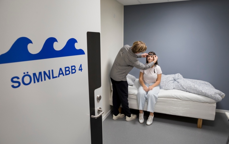 Person sätter någonting runt huvudet på en testdeltagare som sitter på säng. 